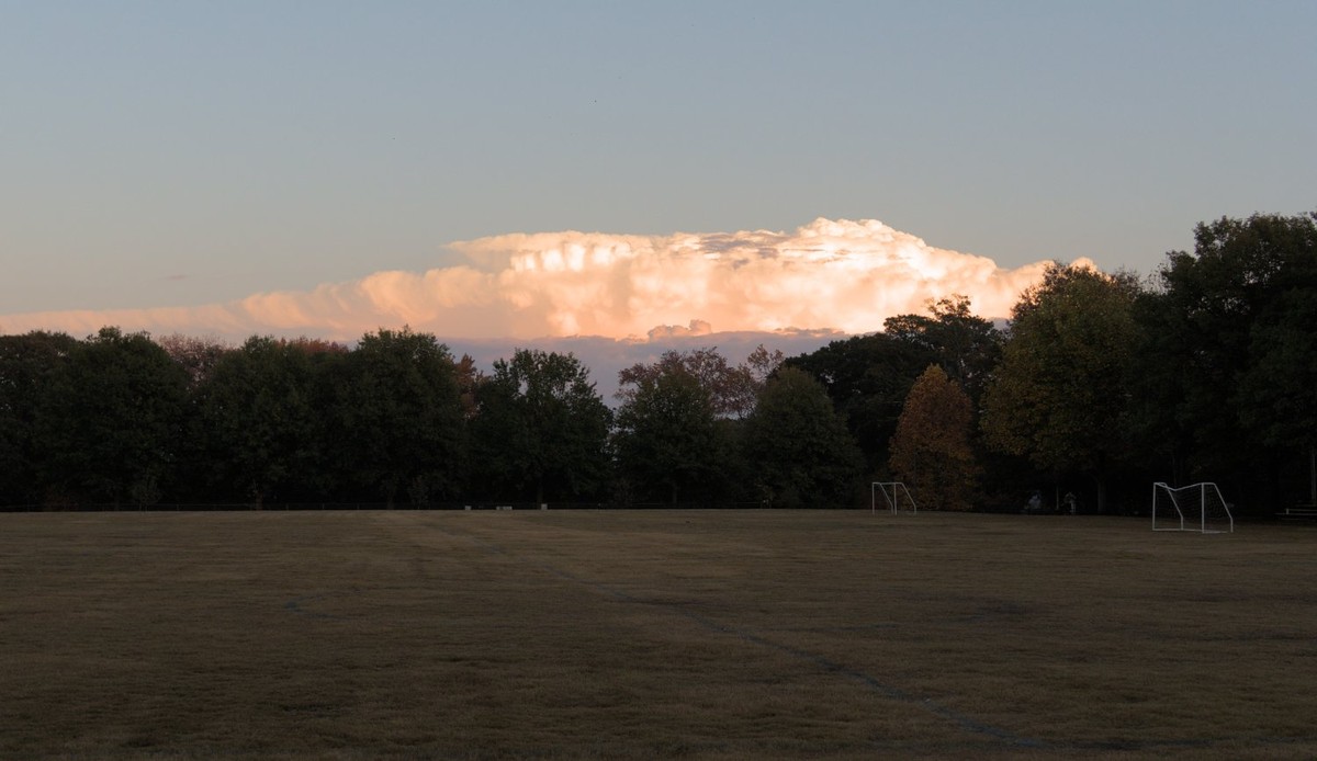 Picture of a park with a large grass soccer field. Trees are along the far end of the park and in the horizon there are some clouds that are bright and orange from the setting sun. 