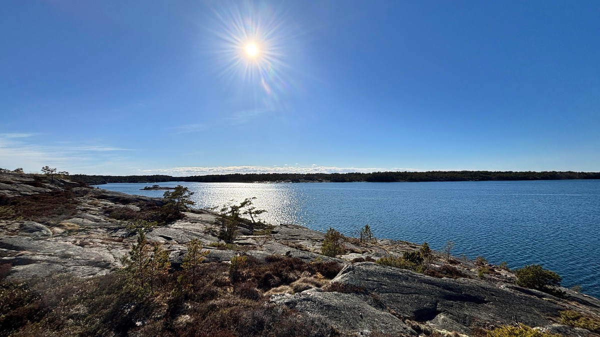 Photo of a high cliff going downwards to the left with water sparkling with sunlight, the forest of another island on the horizon with a bit of clouds beyond it, but mostly blue skies and sun...